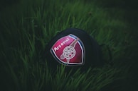 A piece of clothing or accessory, possibly a cap, with the Arsenal football club logo placed on green grass. The logo features prominently in the center with a cannon image on a red and blue shield design.