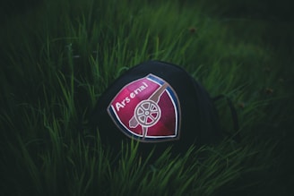 A piece of clothing or accessory, possibly a cap, with the Arsenal football club logo placed on green grass. The logo features prominently in the center with a cannon image on a red and blue shield design.