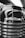 A close-up, black-and-white photograph of the front of a vintage Chevrolet vehicle. The chrome grille features prominently with its shiny, reflective surface. The Chevrolet logo is clearly visible, adding a classic touch. The background includes blurred elements, possibly of a showroom or exhibition space.