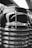 A close-up, black-and-white photograph of the front of a vintage Chevrolet vehicle. The chrome grille features prominently with its shiny, reflective surface. The Chevrolet logo is clearly visible, adding a classic touch. The background includes blurred elements, possibly of a showroom or exhibition space.
