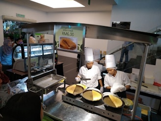 Two chefs wearing white uniforms and tall hats are preparing a dish at a food stall. A sign displays 'Apam Balik RM6.00'. There are several cooking utensils and pans on a metal counter, and a crowd of people stands nearby, suggesting an indoor market or food court setting.