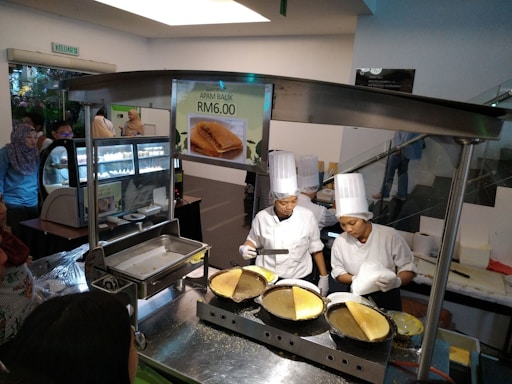 Two chefs wearing white uniforms and tall hats are preparing a dish at a food stall. A sign displays 'Apam Balik RM6.00'. There are several cooking utensils and pans on a metal counter, and a crowd of people stands nearby, suggesting an indoor market or food court setting.