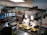 Two chefs wearing white uniforms and tall hats are preparing a dish at a food stall. A sign displays 'Apam Balik RM6.00'. There are several cooking utensils and pans on a metal counter, and a crowd of people stands nearby, suggesting an indoor market or food court setting.