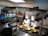 Two chefs wearing white uniforms and tall hats are preparing a dish at a food stall. A sign displays 'Apam Balik RM6.00'. There are several cooking utensils and pans on a metal counter, and a crowd of people stands nearby, suggesting an indoor market or food court setting.