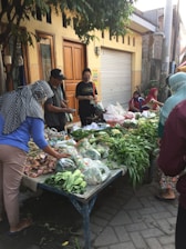 A street market scene with several people gathered around a table covered with various fresh vegetables. The table is set up on a paved sidewalk in front of building entrances. Shoppers engage in selecting produce, including leafy greens and other vegetables, which are displayed in plastic bags. A tree provides partial shade over the area.