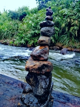 A stack of rounded stones is carefully balanced on a larger rock by the edge of a flowing river. The surrounding environment features dense green foliage, suggesting a lush, natural setting.