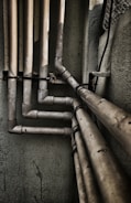 Multiple interconnected pipes run vertically and horizontally along the corner of a slightly worn and grimy wall. The pipes have a weathered and rusty appearance, suggesting they have been in place for some time. Some cables are also visible next to the pipes.