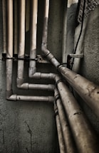 Multiple interconnected pipes run vertically and horizontally along the corner of a slightly worn and grimy wall. The pipes have a weathered and rusty appearance, suggesting they have been in place for some time. Some cables are also visible next to the pipes.
