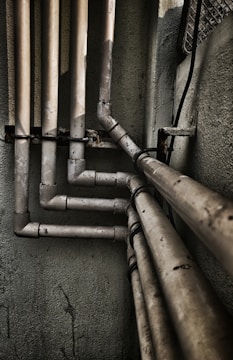 Multiple interconnected pipes run vertically and horizontally along the corner of a slightly worn and grimy wall. The pipes have a weathered and rusty appearance, suggesting they have been in place for some time. Some cables are also visible next to the pipes.
