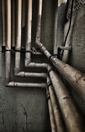 Multiple interconnected pipes run vertically and horizontally along the corner of a slightly worn and grimy wall. The pipes have a weathered and rusty appearance, suggesting they have been in place for some time. Some cables are also visible next to the pipes.