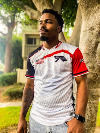 A person is standing outdoors, wearing a sports jersey with red, white, and black design elements, including an American flag patch. They have short hair and a beard, a tattoo on one arm, and are wearing a smartwatch. The background features trees and a blurred house façade, giving a sense of a suburban or park setting.