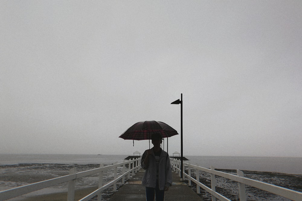 日中、傘をさして桟橋に立つ黒いジャケットとズボンを着た人物