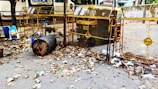 A street scene with a large metal trash bin overflowing with garbage, surrounded by dried leaves. There are blue and black plastic bags, along with scattered litter around the bin. The area is enclosed by a yellow barricade with some signs attached, and an auto rickshaw is parked behind the barricade. The surrounding environment appears to be a residential or commercial area with some trees and buildings in the background.