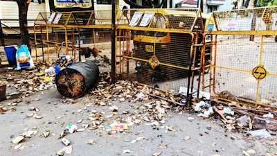 A street scene with a large metal trash bin overflowing with garbage, surrounded by dried leaves. There are blue and black plastic bags, along with scattered litter around the bin. The area is enclosed by a yellow barricade with some signs attached, and an auto rickshaw is parked behind the barricade. The surrounding environment appears to be a residential or commercial area with some trees and buildings in the background.