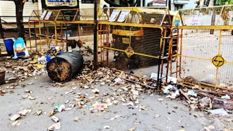 A street scene with a large metal trash bin overflowing with garbage, surrounded by dried leaves. There are blue and black plastic bags, along with scattered litter around the bin. The area is enclosed by a yellow barricade with some signs attached, and an auto rickshaw is parked behind the barricade. The surrounding environment appears to be a residential or commercial area with some trees and buildings in the background.