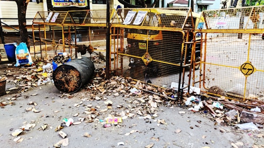 A street scene with a large metal trash bin overflowing with garbage, surrounded by dried leaves. There are blue and black plastic bags, along with scattered litter around the bin. The area is enclosed by a yellow barricade with some signs attached, and an auto rickshaw is parked behind the barricade. The surrounding environment appears to be a residential or commercial area with some trees and buildings in the background.