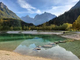 A serene mountain landscape with a clear, calm lake in the foreground. The water reflects the surrounding mountains and forest, adding a sense of tranquility. The mountains are capped with snow, and dense forest lines the shoreline, displaying autumnal colors. Stepping stones lead into the lake, inviting a sense of exploration.