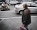 An elderly person stands on a sidewalk holding a red bag filled with containers. Several cars are visible on the road, including a silver sedan and a black SUV. A person on a scooter is blurred, crossing in the opposite direction.