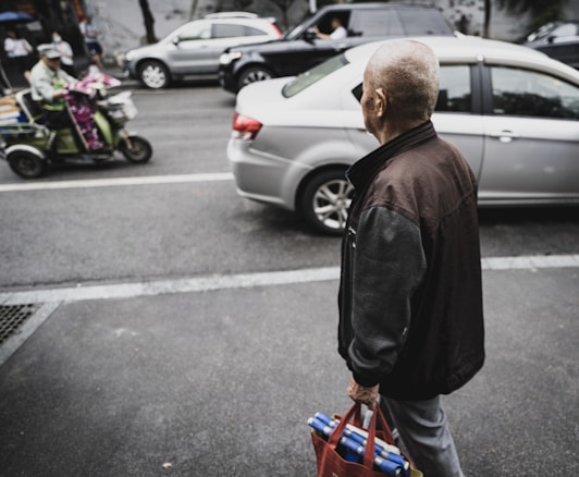 An elderly person stands on a sidewalk holding a red bag filled with containers. Several cars are visible on the road, including a silver sedan and a black SUV. A person on a scooter is blurred, crossing in the opposite direction.