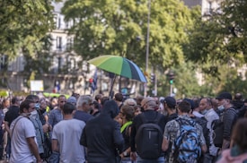 A large group of people gathered on a street, surrounded by tall trees and urban buildings. Some individuals are wearing masks, and one person is holding a green and black umbrella. The scene suggests a public event or protest with a mix of casual and serious expressions.