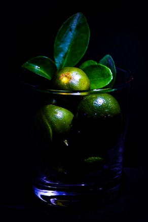 A glass filled with green limes and vivid green leaves, set against a dark background. The lighting accentuates the glossy texture of the limes and the smooth surface of the leaves, creating a strong contrast.