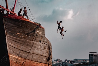Several young individuals are on the deck of a large, weathered wooden ship, with two of them captured mid-air as they jump off the side. The sky is partly cloudy, and urban buildings can be seen in the background, giving a sense of adventure and heedless joy.