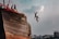Several young individuals are on the deck of a large, weathered wooden ship, with two of them captured mid-air as they jump off the side. The sky is partly cloudy, and urban buildings can be seen in the background, giving a sense of adventure and heedless joy.