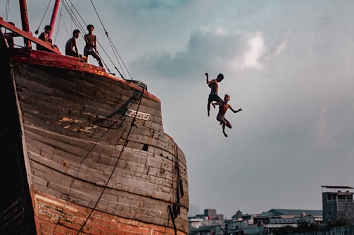 Several young individuals are on the deck of a large, weathered wooden ship, with two of them captured mid-air as they jump off the side. The sky is partly cloudy, and urban buildings can be seen in the background, giving a sense of adventure and heedless joy.