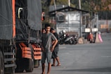 A man stands next to a large truck, which has a black tarp covering its side. Several other trucks appear lined up, and motorcycles are parked in the background. The area looks like an industrial or loading zone with concrete and metal structures.