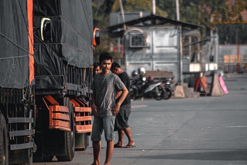 A man stands next to a large truck, which has a black tarp covering its side. Several other trucks appear lined up, and motorcycles are parked in the background. The area looks like an industrial or loading zone with concrete and metal structures.