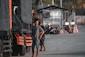 A man stands next to a large truck, which has a black tarp covering its side. Several other trucks appear lined up, and motorcycles are parked in the background. The area looks like an industrial or loading zone with concrete and metal structures.