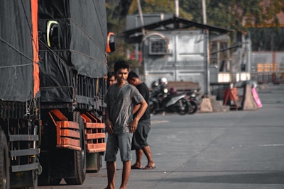 A man stands next to a large truck, which has a black tarp covering its side. Several other trucks appear lined up, and motorcycles are parked in the background. The area looks like an industrial or loading zone with concrete and metal structures.