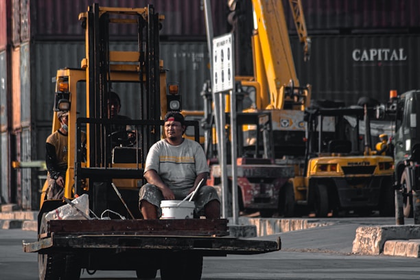A man sits on the front of a forklift holding a bucket in an industrial area filled with shipping containers and heavy machinery. The background shows multiple cranes and vehicles, indicating a busy shipping or construction site.