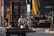 A man sits on the front of a forklift holding a bucket in an industrial area filled with shipping containers and heavy machinery. The background shows multiple cranes and vehicles, indicating a busy shipping or construction site.