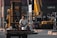 A man sits on the front of a forklift holding a bucket in an industrial area filled with shipping containers and heavy machinery. The background shows multiple cranes and vehicles, indicating a busy shipping or construction site.