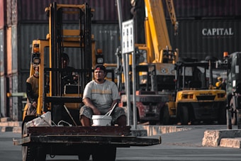 A man sits on the front of a forklift holding a bucket in an industrial area filled with shipping containers and heavy machinery. The background shows multiple cranes and vehicles, indicating a busy shipping or construction site.