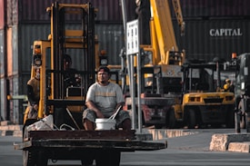 A man sits on the front of a forklift holding a bucket in an industrial area filled with shipping containers and heavy machinery. The background shows multiple cranes and vehicles, indicating a busy shipping or construction site.