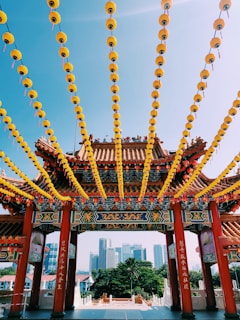 A traditional Chinese temple gate adorned with intricate designs and vibrant colors is in the foreground. Numerous yellow lanterns hang in rows across the sky, creating a festive atmosphere. In the background, modern skyscrapers rise against a clear blue sky, juxtaposing the ancient with the contemporary. Lush greenery surrounds the temple, highlighting its cultural significance.