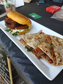 A white rectangular plate holds a hamburger on the left and a stack of quesadillas on the right. The hamburger features a thick patty, lettuce, and a bun with a golden hue. The quesadillas are filled with meat, cheese, and some spinach, showcasing a crispy, grilled exterior. The setting includes a dark table surface with various objects like a green card, a transparent cup with red contents, and a red packet in the background.