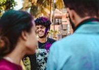 A young man with curly hair is laughing, his face and clothes covered in colorful powder. Two people are in the foreground with their backs to the camera, partially obstructing the view. Palm trees and a building are visible in the background.
