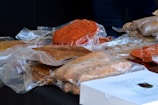 A variety of packaged meats are displayed on a black surface. The meats are vacuum-sealed in transparent plastic wrapping, showcasing different types such as a piece with a reddish hue and others with brownish tones. A white box with printed text is partially visible in the foreground.