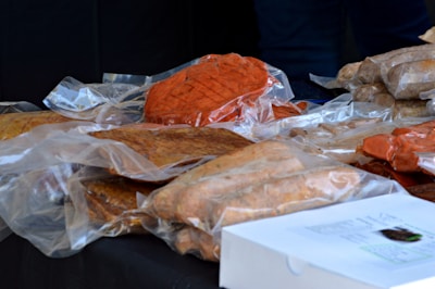A variety of packaged meats are displayed on a black surface. The meats are vacuum-sealed in transparent plastic wrapping, showcasing different types such as a piece with a reddish hue and others with brownish tones. A white box with printed text is partially visible in the foreground.