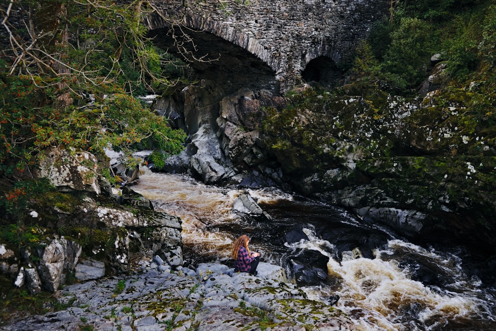 woman in red jacket sitting on rock near river during daytime