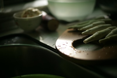 A dimly lit kitchen scene with a wooden cutting board in the foreground. Several okra pods are arranged on the board, glistening with water droplets. In the blurry background, a small bowl contains a scoop of some substance, possibly ice cream or butter, and an unidentifiable root vegetable is partially visible.