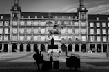 A black and white image featuring a large, historic plaza with an equestrian statue at the center. A silhouetted artist sits in the foreground, seemingly sketching or painting. The background consists of an ornate, multi-story building with arches, balconies, and decorative elements. Several people are walking around the plaza.