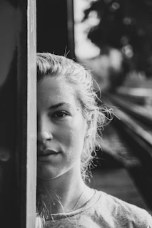 A black and white portrait of a person partially obscured by a vertical surface. The focus is on the face, capturing an introspective or contemplative expression. The hair is pulled back, and the background includes blurred train tracks leading into the distance.