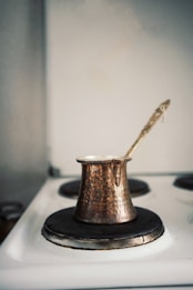 A traditional, hammered copper Turkish coffee pot sits on an electric stove. The pot has a long, ornate handle extending from the top. The stove is white with black burners, showing an old-fashioned style. The overall setting suggests a cozy, rustic kitchen atmosphere.