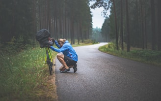 A cyclist in a blue jacket is crouched down on the side of a wet road, attending to a camera mounted on a yellow touring bike with a large black bag on the back. The road is surrounded by a forest of tall pine trees, creating a serene and somewhat lonely atmosphere.
