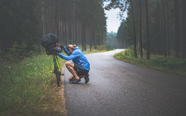 A cyclist in a blue jacket is crouched down on the side of a wet road, attending to a camera mounted on a yellow touring bike with a large black bag on the back. The road is surrounded by a forest of tall pine trees, creating a serene and somewhat lonely atmosphere.