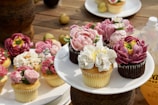 An assortment of cupcakes decorated with intricate, flower-shaped frosting, displayed on a wooden surface. The cupcakes feature a variety of colors, including pink, white, and purple, and are arranged on a white plate. Additional cupcakes are scattered around the main display, and there are some small fruits and a bottle in the background.